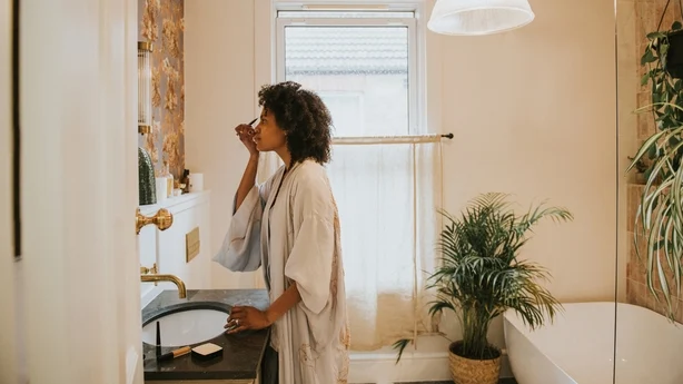 En smuk sort kvinde står i et stilfuldt badeværelse i en lang, silkeblød kjortel og lægger omhyggeligt sin make-up.